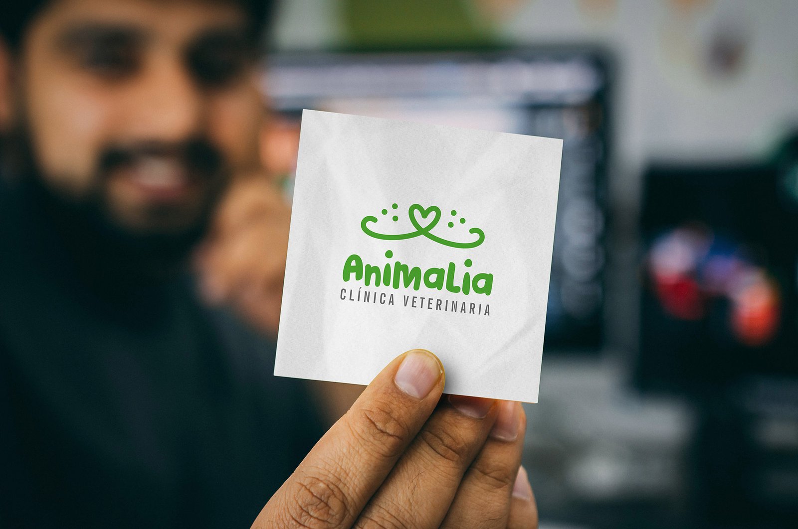 Dedos sosteniendo un papel blanco, que tiene el Logo Animalia - Clínica Veterinaria. Al fondo se ve muy desenfocado, el hombre que sostiene el pequeño papel, con ambiente de trabajo.