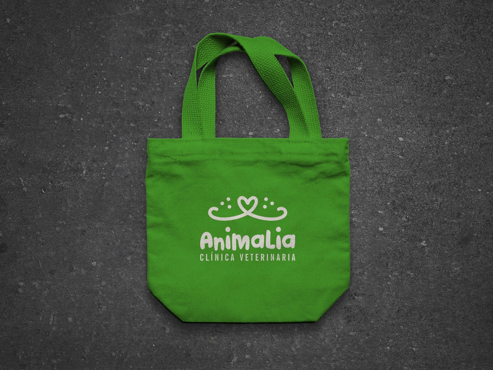 Logo Animalia - Clínica Veterinaria en blanco, montado sobre una pequeña bolsita de tela color verde, con agarraderas. Fondo gris oscuro con textura de concreto.